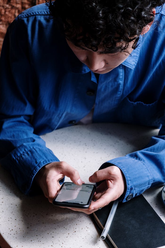 about-01 A young man in a blue shirt uses his smartphone in a cozy café, focusing on typing messages.